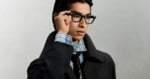 A young man with dark hair wears black-rimmed glasses, a light blue striped shirt, a dark tie, and a dark coat, with his hand resting on the frame of his glasses.
