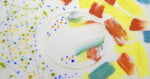 A circular glass pane sits on a white surface, featuring colorful paint splatters on the left and bold, textured brushstrokes in yellow, orange, and blue on the right.