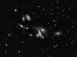 A deep-space view showing a dense cluster of various spiral and elliptical galaxies scattered across a dark, star-filled background.