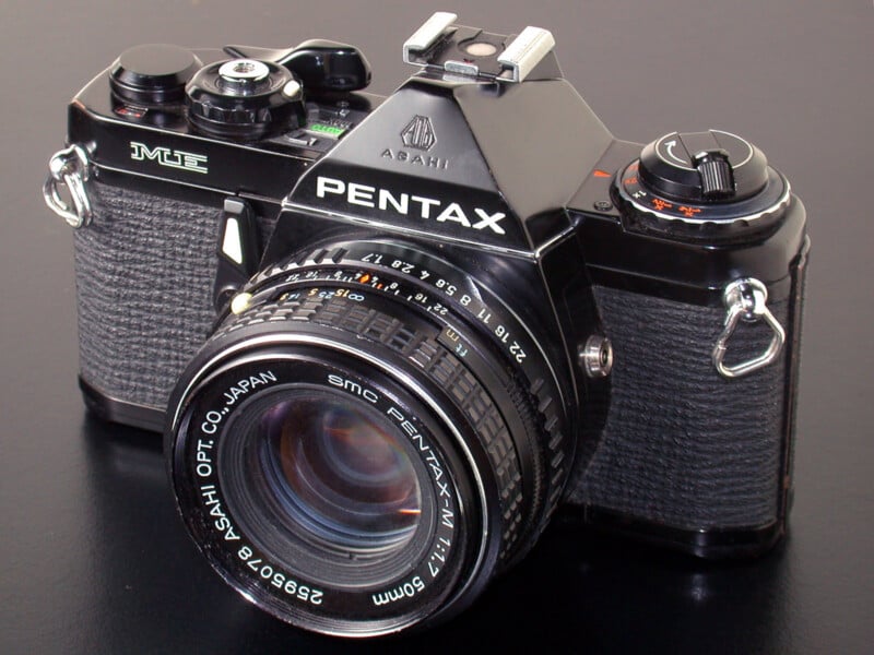 A black Pentax ME 35mm film camera with an SMC Pentax-M 50mm f/1.7 lens, positioned on a dark, reflective surface.