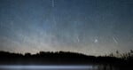 A night sky filled with stars and several bright meteor streaks above a dark, silhouetted forest and a misty lake.
