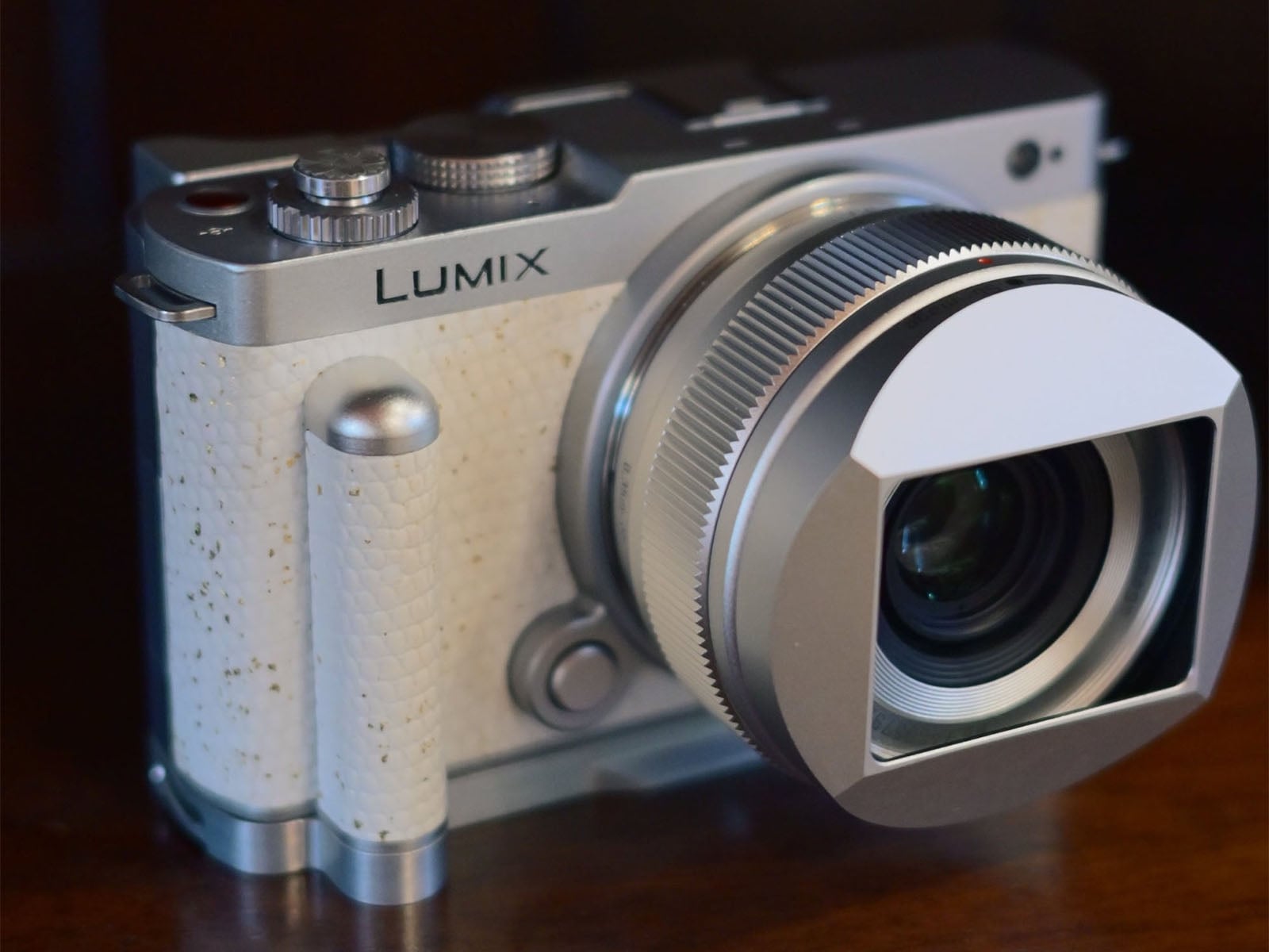 A silver Lumix digital camera with a white textured grip sits on a dark wooden surface, featuring a lens with a square-shaped hood.