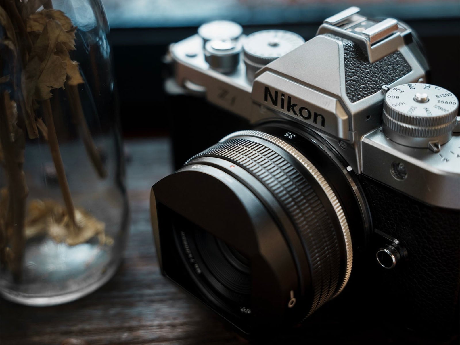 A silver and black Nikon Zf mirrorless camera with a prime lens sits on a wooden surface next to a glass vase containing dried plants.