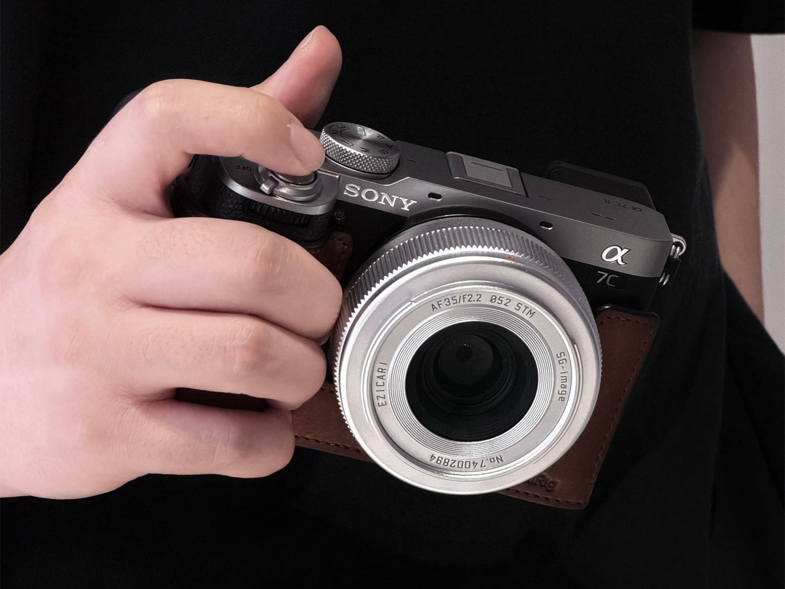 A person's hand holds a silver and black Sony Alpha 7C II camera equipped with an Ezicari 35mm f/2.2 lens and a brown leather case.