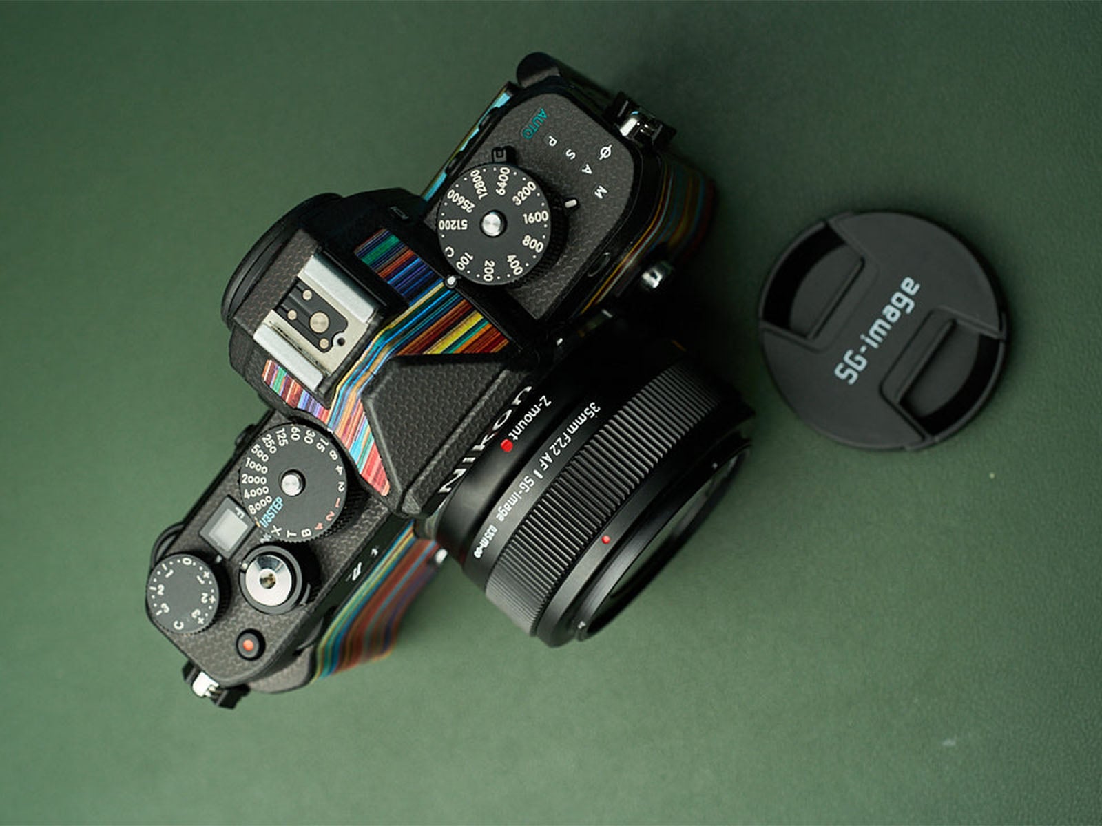 A black mirrorless camera with a colorful, striped skin sits on a green surface next to a detached lens cap labeled "SG-image.