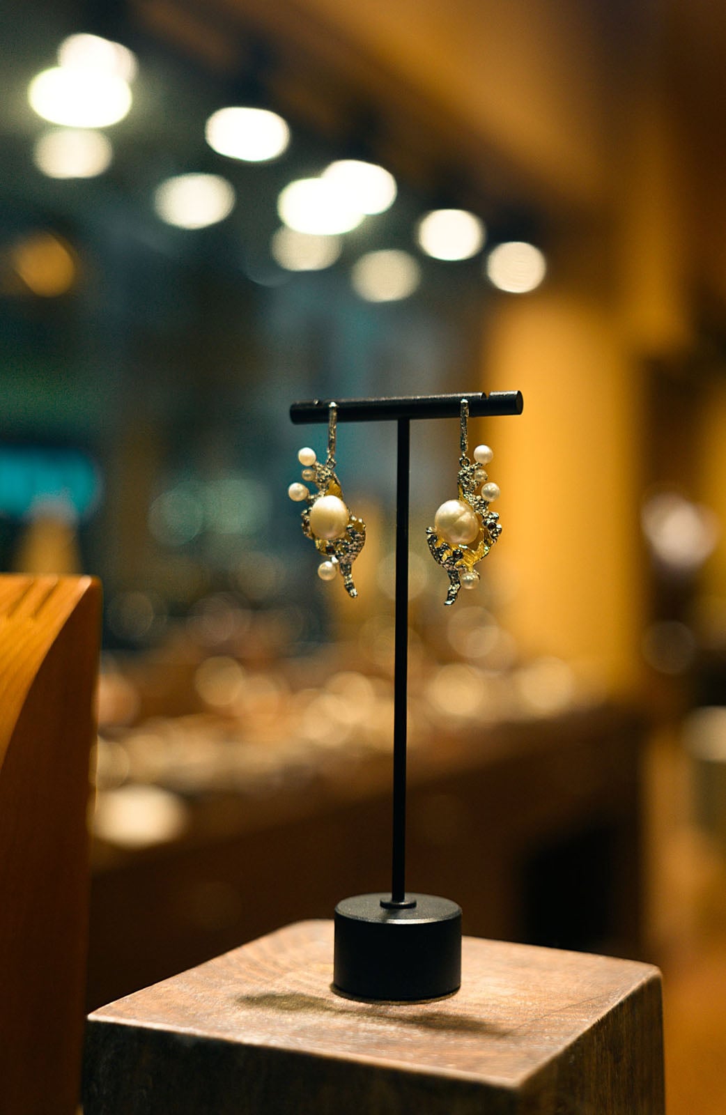 A pair of ornate, pearl-accented earrings hangs from a black T-shaped jewelry stand on a wooden block against a blurred, warm-toned background.