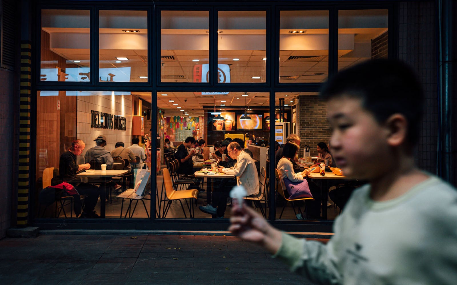 A young boy walks past a brightly lit restaurant window where several people are seated at tables eating.