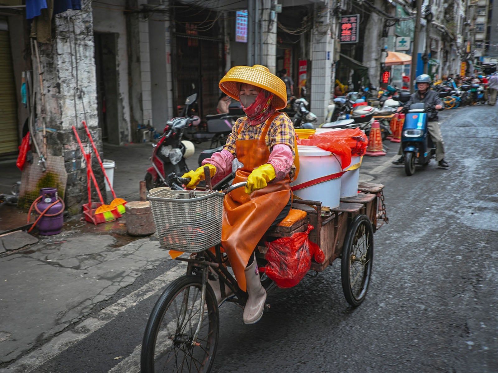 A person wearing a yellow hat, face mask, and orange apron rides a three-wheeled cargo bicycle carrying large white buckets down a city street.