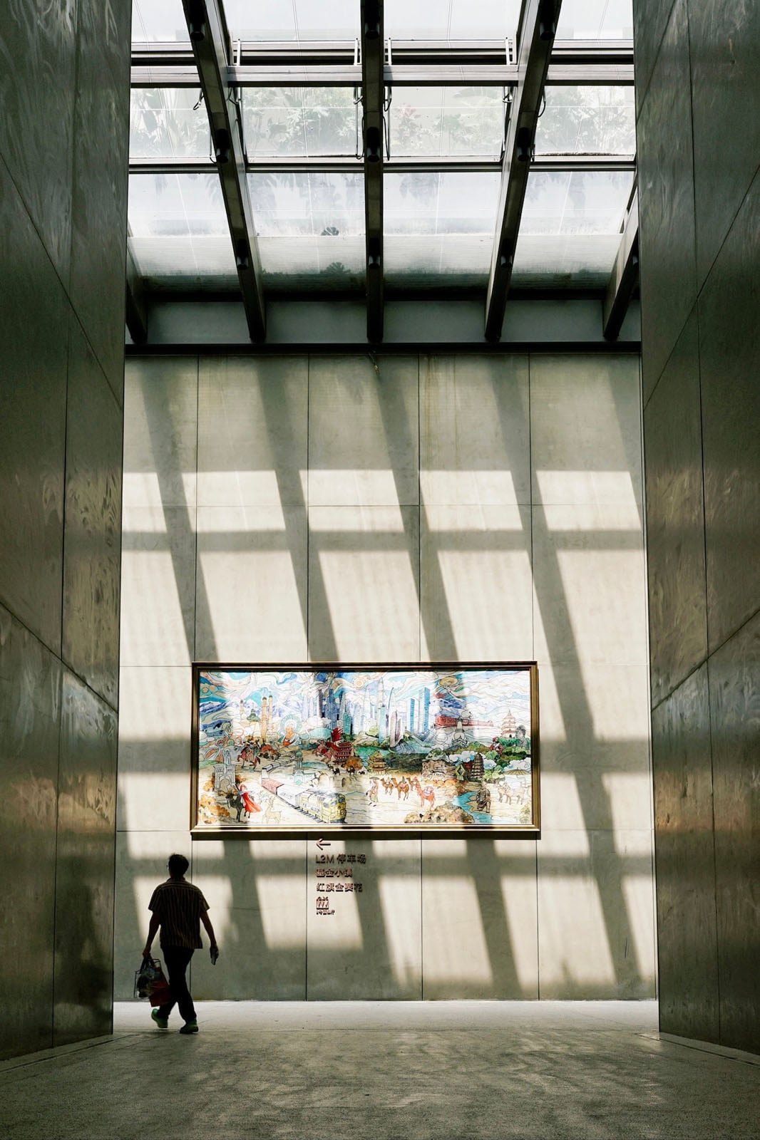 A person walks through a sunlit corridor toward a large, framed mural depicting a landscape with buildings and figures on a concrete wall.