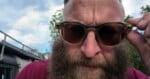 A close-up shot of a man with a thick beard wearing translucent-framed sunglasses, holding the frame with his hand.