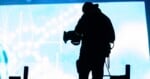A silhouetted videographer stands in front of a bright, glowing blue screen while operating a professional video camera.