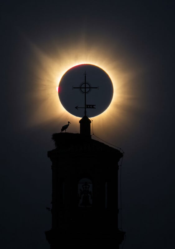 A solar eclipse appears directly behind the cross-topped spire of a bell tower, where a stork stands in a nest.