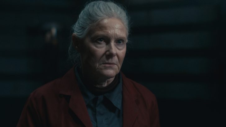 Dead ResetElderly woman with gray hair looks serious in a dimly lit setting, wearing a dark red jacket.