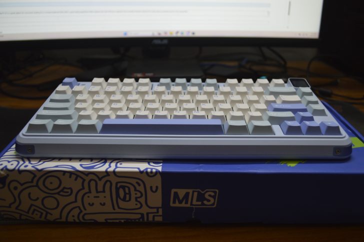 Blue and white mechanical keyboard on a decorated box, monitor in the background.