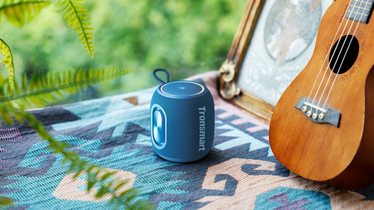 Blue portable Bluetooth speaker on a colorful cloth near a ukulele and a picture frame.