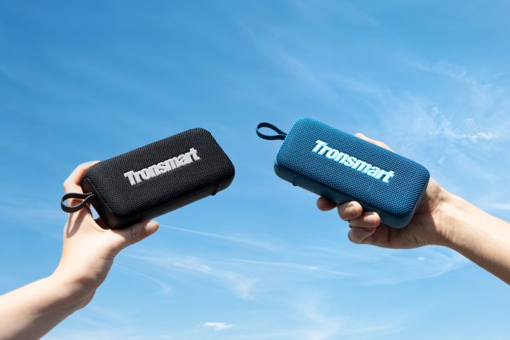 Two hands holding black and blue Tronsmart speakers against a clear blue sky.