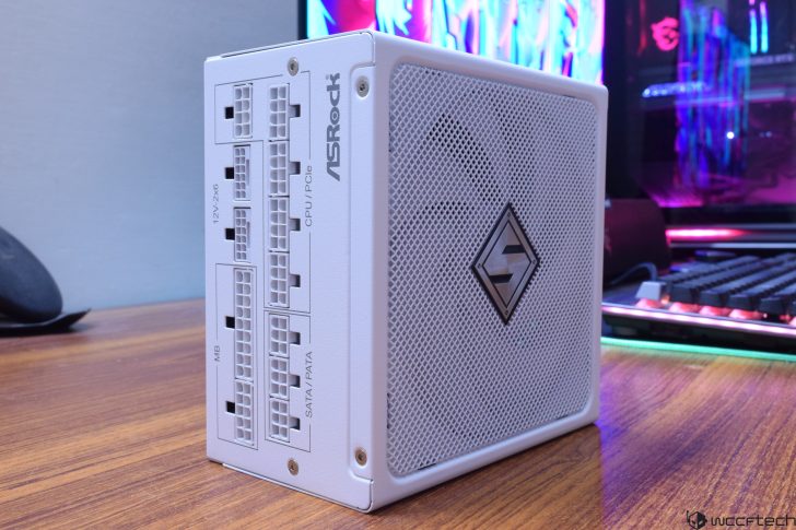 White ASRock computer power supply on wooden desk with colorful gaming setup in the background.