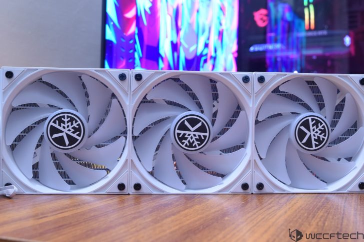A close-up of three Snowflake-branded computer cooling fans arranged side by side on a wooden surface, with a colorful monitor screen in the background.