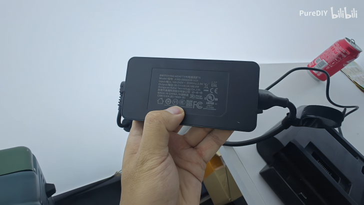 Person holding a black power adapter with cables and labels, desk and soda can in background.