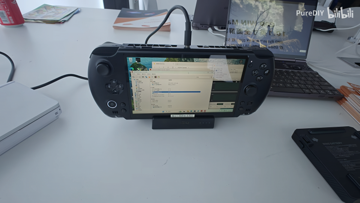 Handheld gaming console on a desk connected to a monitor, laptop in the background.
