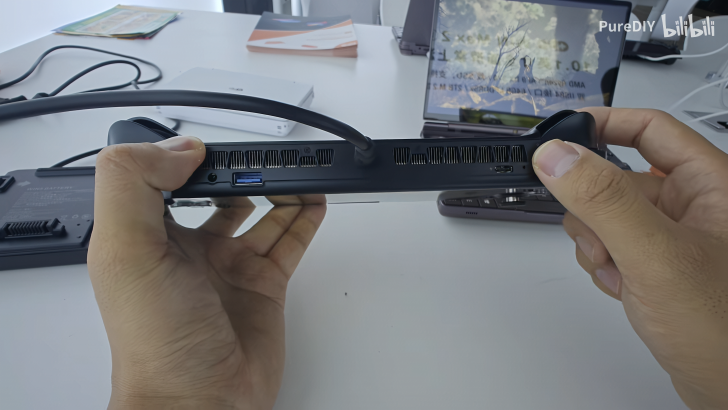 Person holding black gaming console with visible USB ports and vents on a desk.