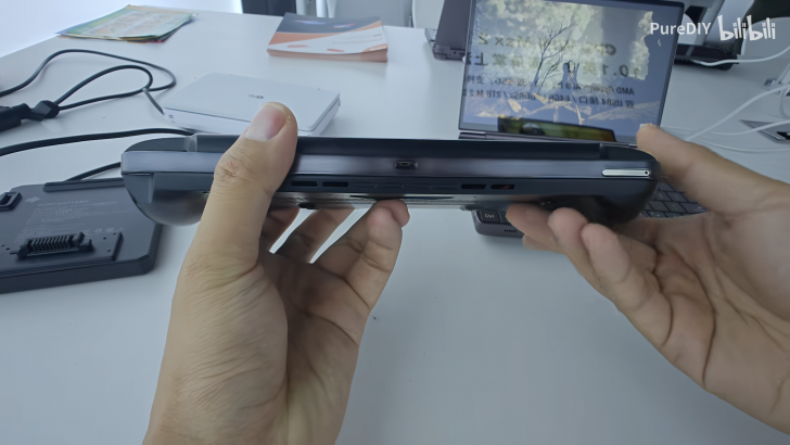 Hands holding a handheld gaming device, laptop in background on a desk.