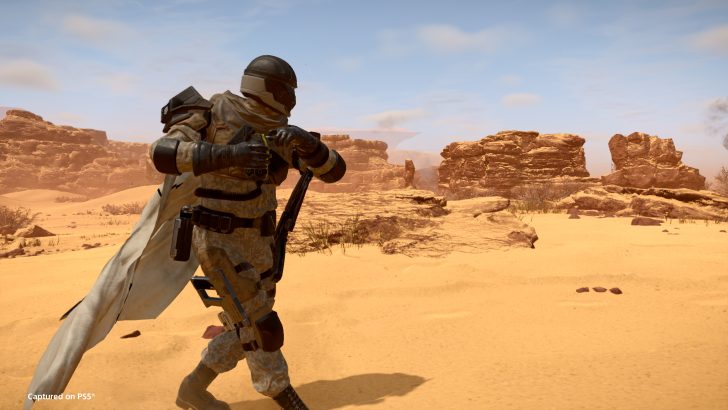 Soldier in desert gear with rifle, exploring rocky landscape under bright blue sky.
