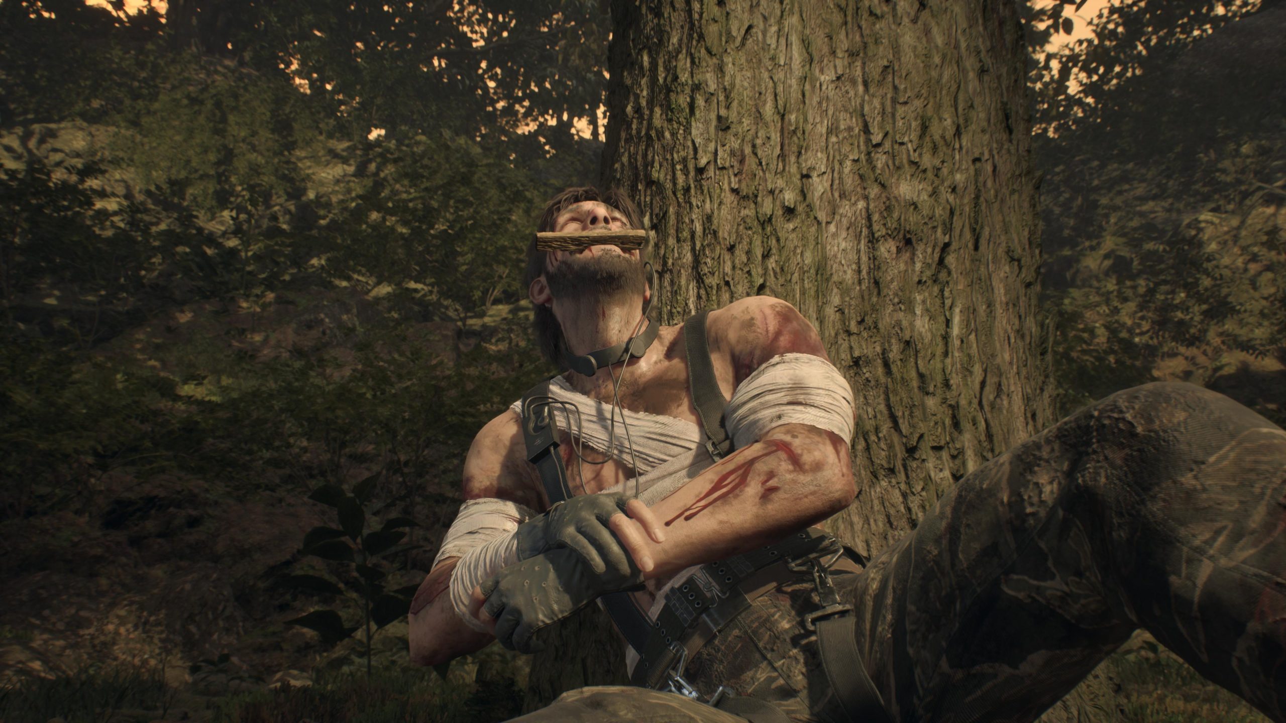 Injured man in forest resting against tree, bandaged arms, biting branch for pain relief.