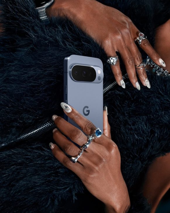 Sleek smartphone with dual cameras held in hands, adorned with rings, over a dark fur coat.