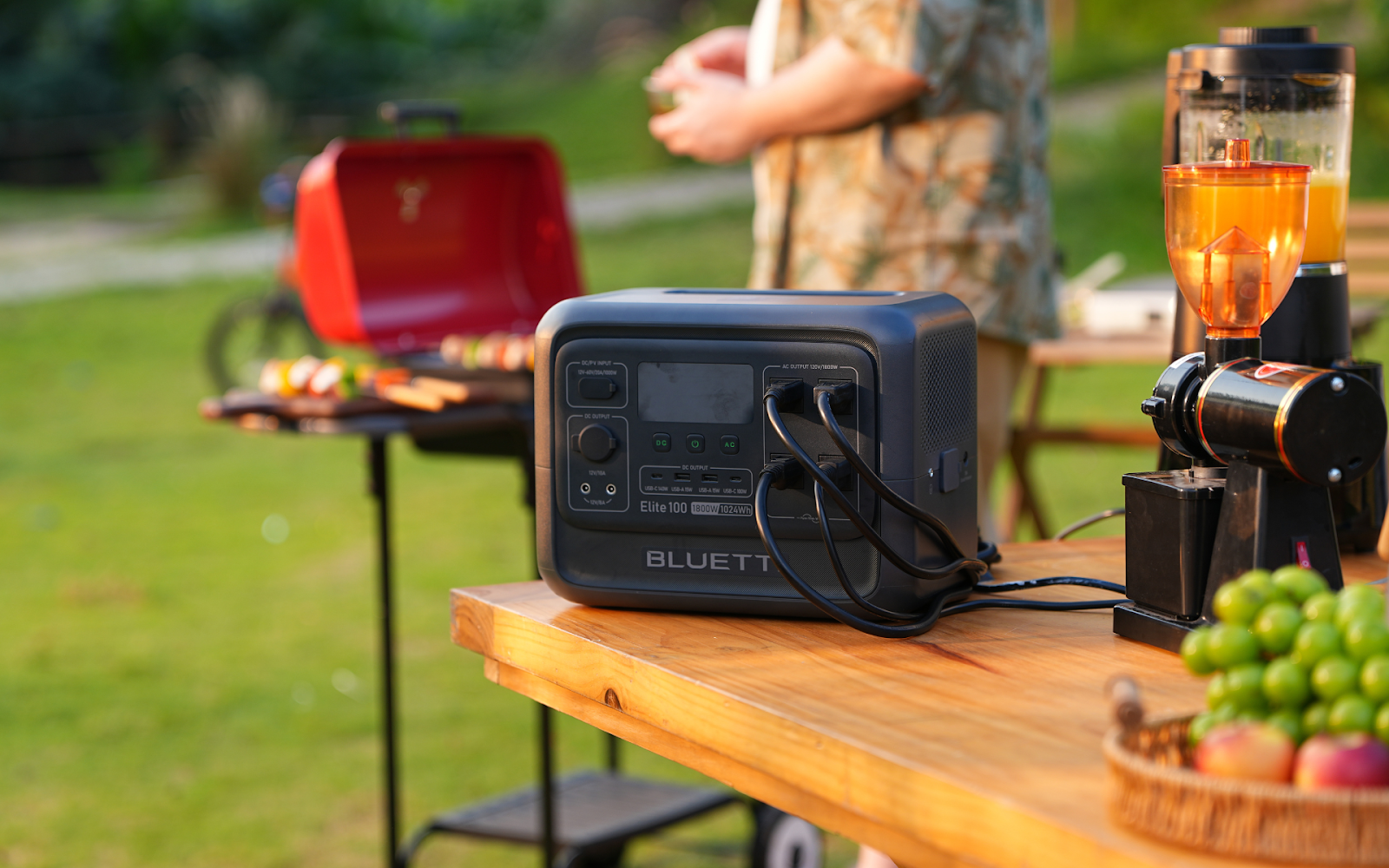 Bluetti Elite 100 V2 portable power station on table at backyard BBQ with juicer and grill.