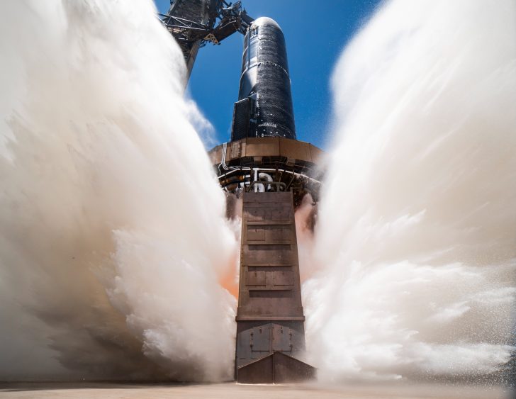 Starship rocket during test fire, releasing powerful exhaust clouds against a clear blue sky, captured from below.