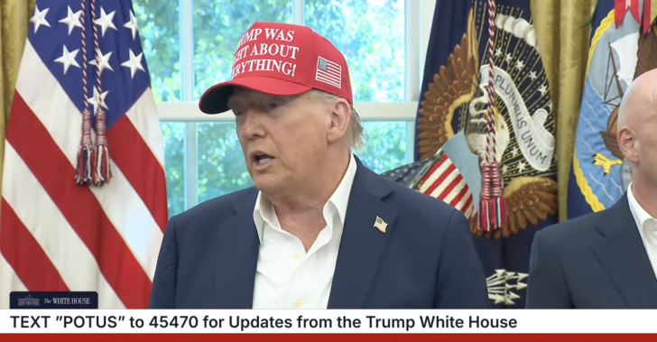 Man in red hat speaking at a press event with flags in the background.