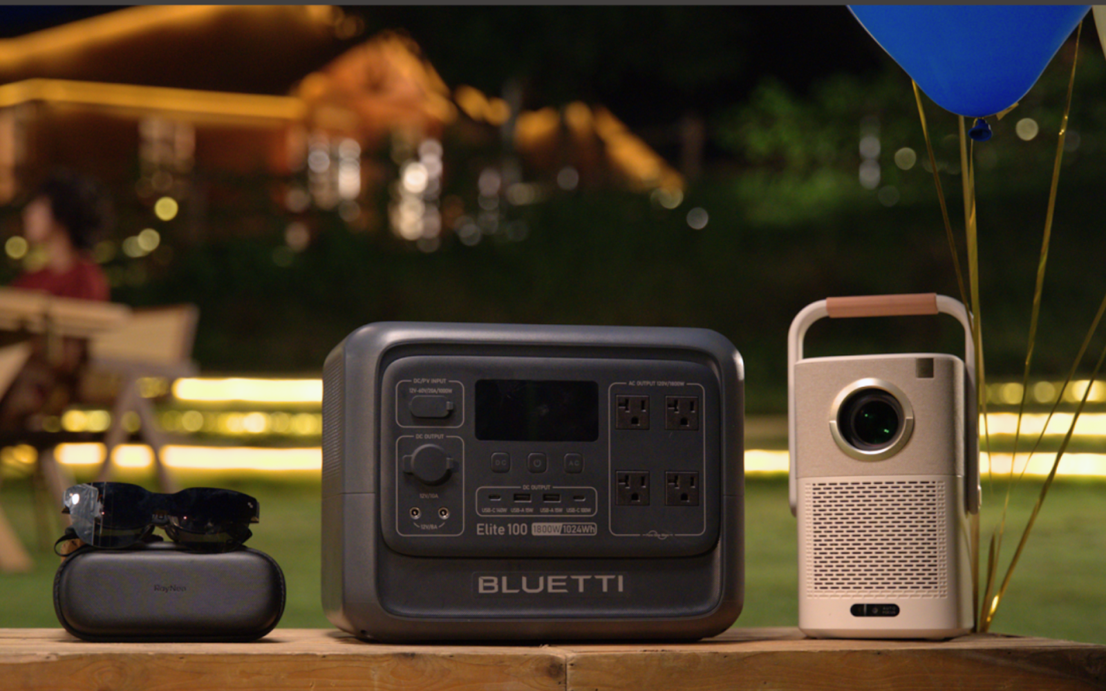 Bluetti portable power station, Yaber projector, and RayNeo AR sunglasses on wooden table outdoors at night.