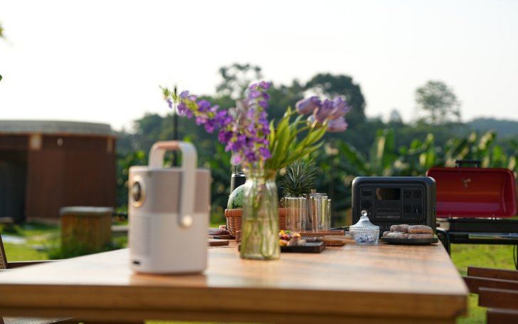 Outdoor picnic table with flowers, Yaber projector, Bluetti power station, and grill in a garden setting.