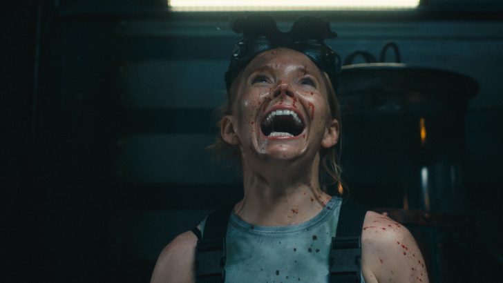 Person with blood-splattered face, wearing night vision goggles, looking up in a dimly lit room.
