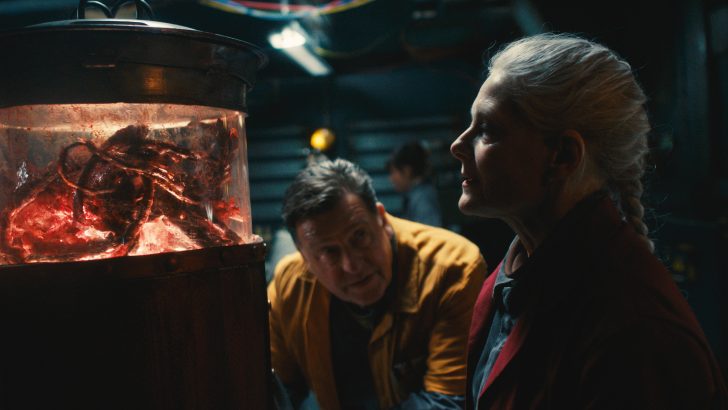 Two people observe a glowing container with an alien-like creature inside in a dimly lit room.