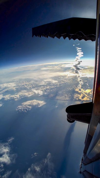 View from aircraft window showing smoke trail over a coastline and ocean.