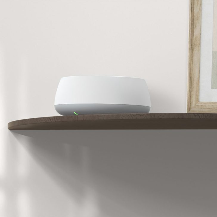 A white Nest Wi-Fi router with a glowing green light sits on a wooden shelf against a light-colored wall.