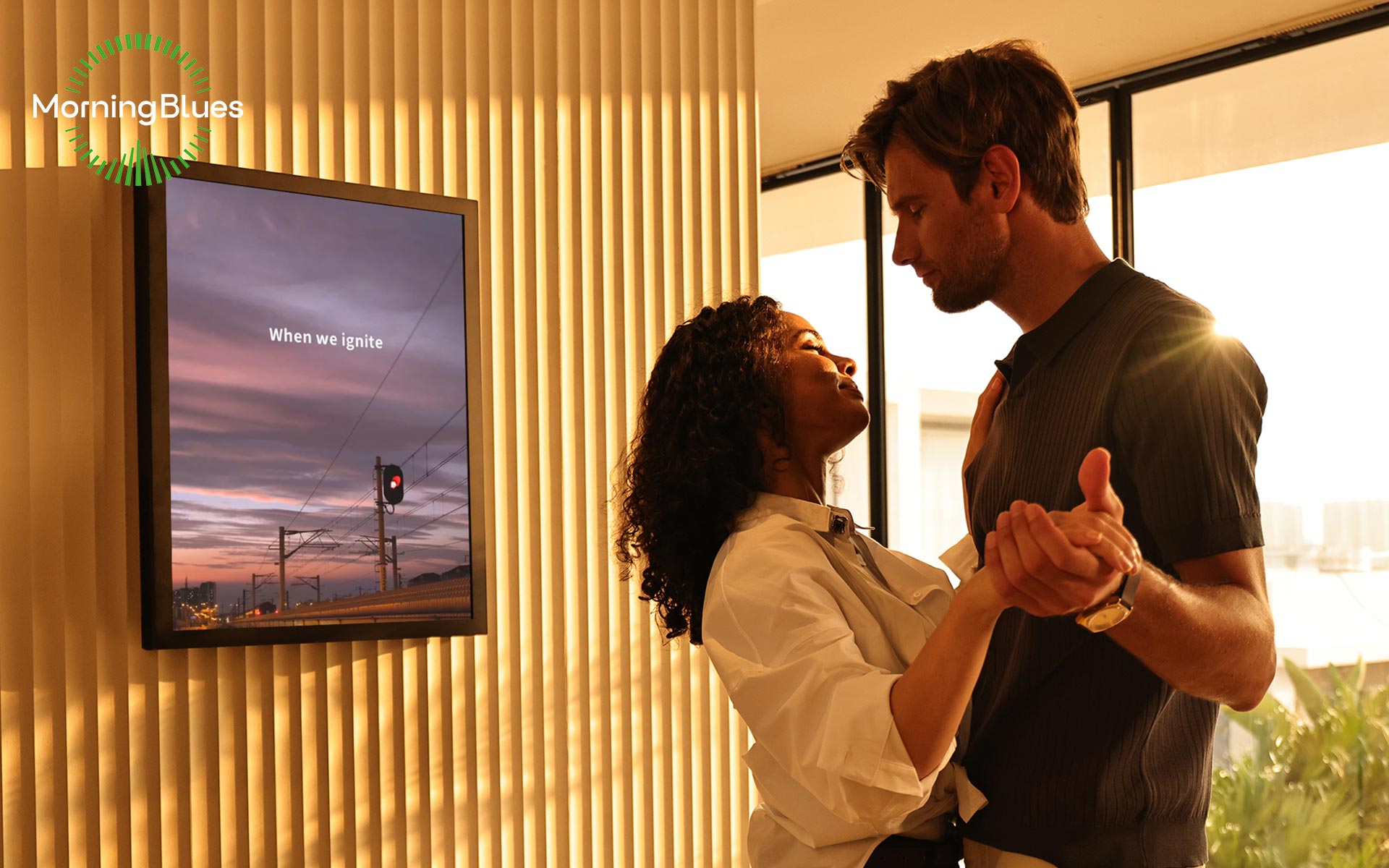 A couple embraces in front of a screen displaying the text 'When we ignite' with a logo that says 'MorningBlues' in a room illuminated by warm sunlight.