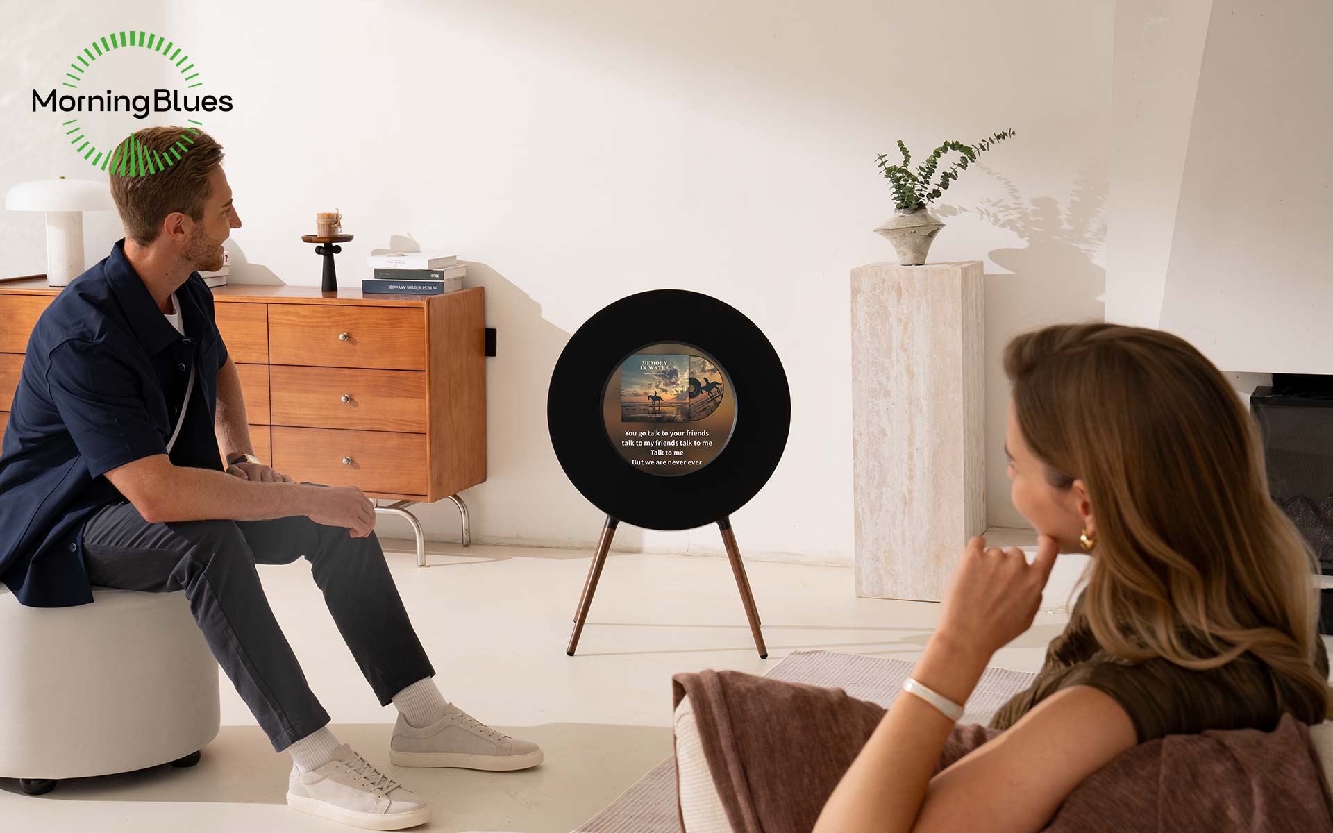 A man and woman sit in a modern living room with a black circular speaker displaying song lyrics, and 'MorningBlues' logo in the corner.
