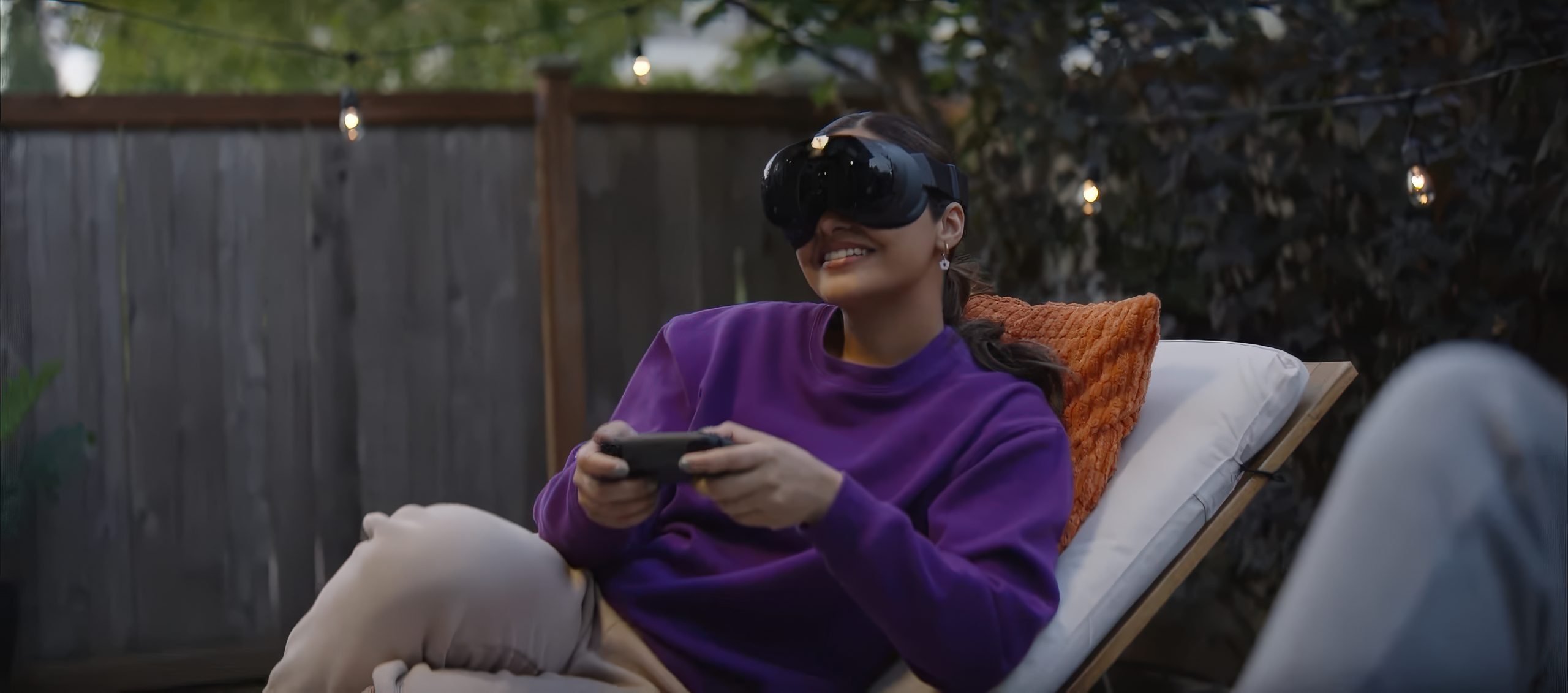 A person wearing Steam Frame headset sits outdoors on a lounge chair, holding a gaming controller and smiling.