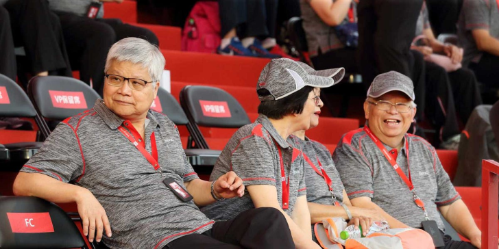 Three people wearing gray and red uniforms with 'BYD' written on them sit on black chairs labeled 'VIP专座' and 'FC' at an indoor event.