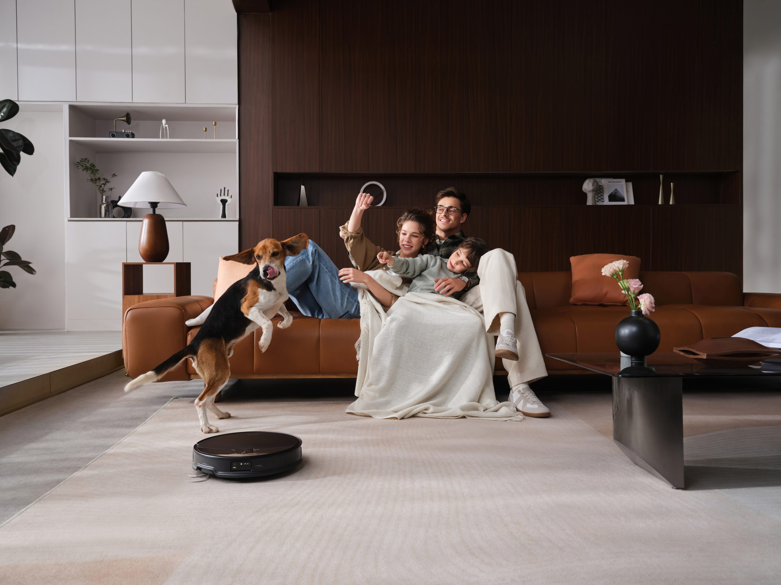A family sits together on a brown leather sofa, while a beagle dog jumps nearby and an unbranded robot vacuum cleans the floor in a modern living room.
