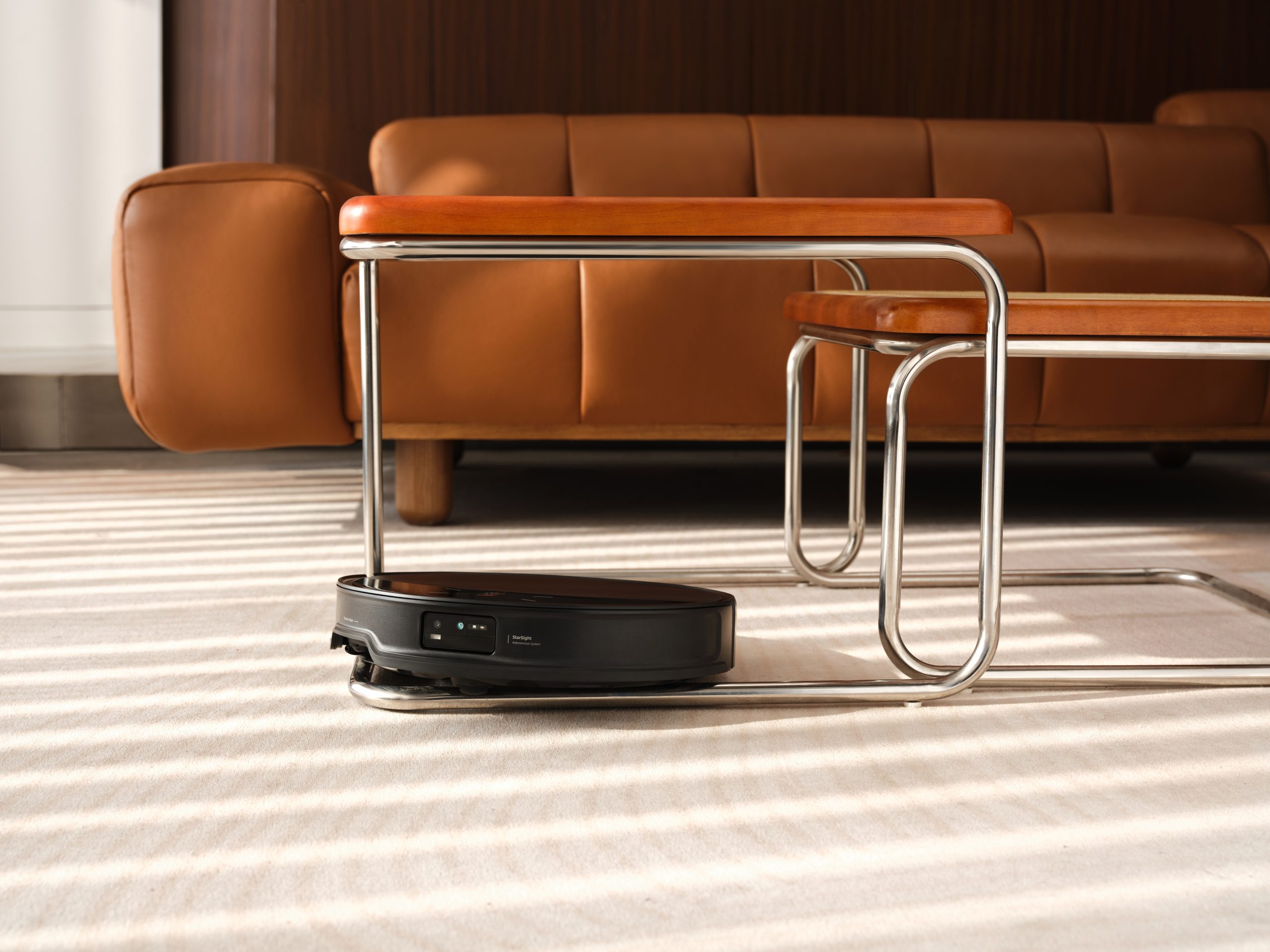 A Roborock S7 Max Ultra robot vacuum cleaner navigates under a table near a brown leather sofa.