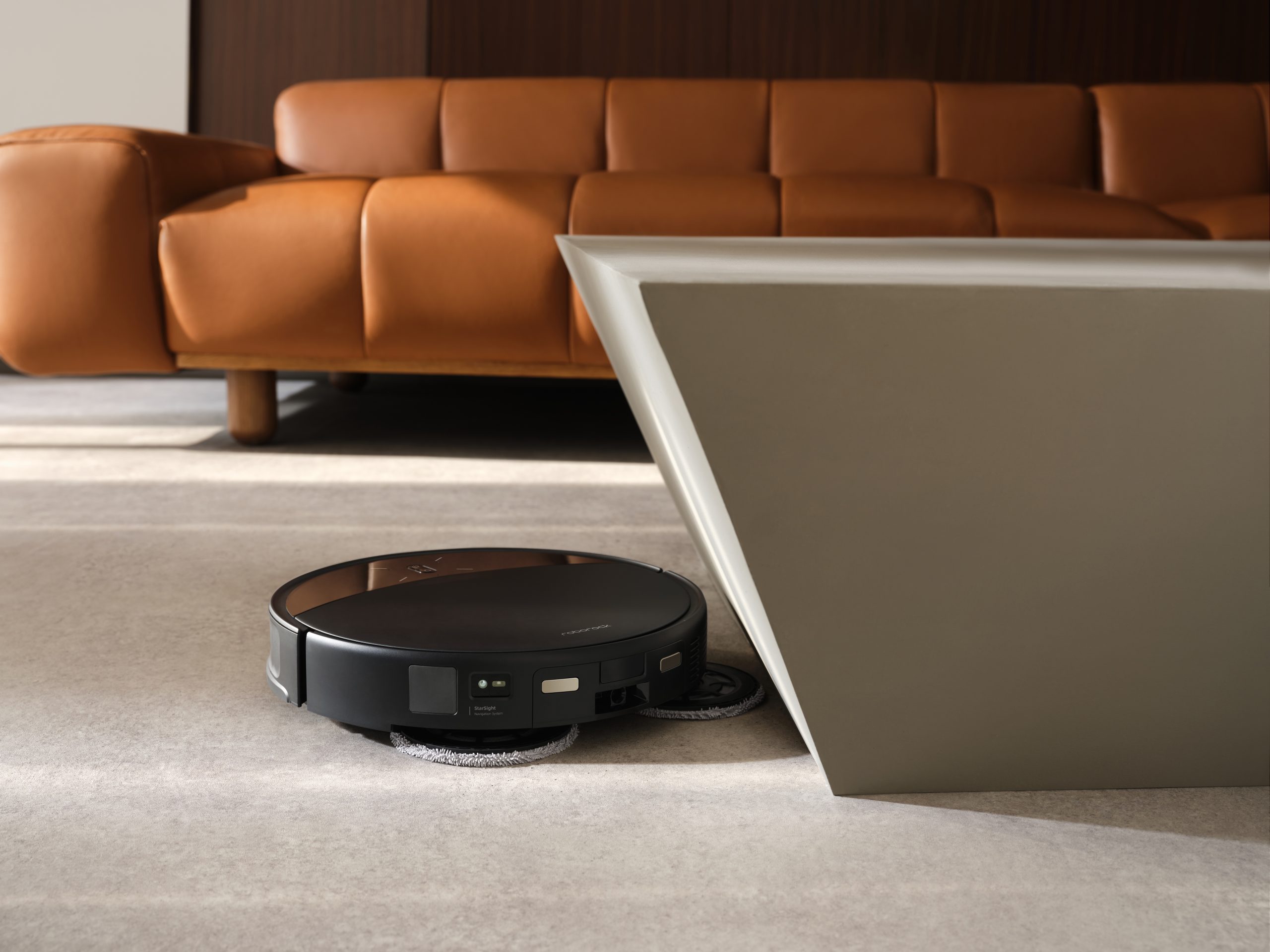 A Roborock robot vacuum cleaner operating on a carpet in a modern living room setting with a brown leather couch and a geometric table.