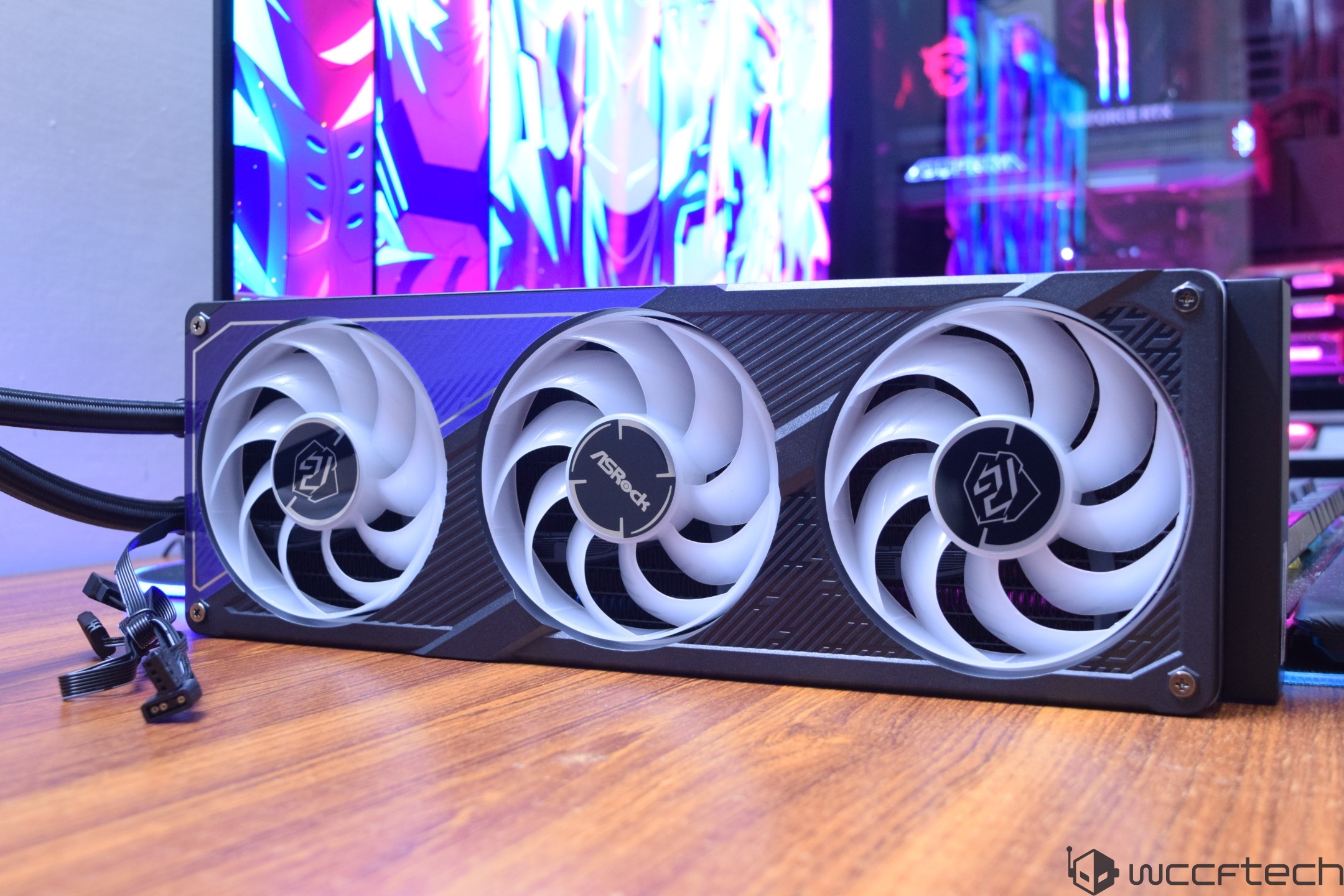An ASRock liquid cooler with three fans on a wooden surface, with a colorful RGB-lit computer setup in the background.