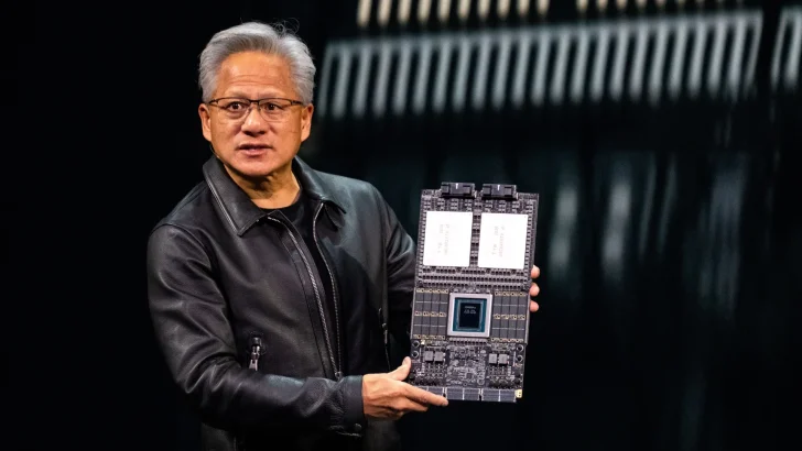 A presenter in a black leather jacket holds a large computing board with visible components and circuitry.