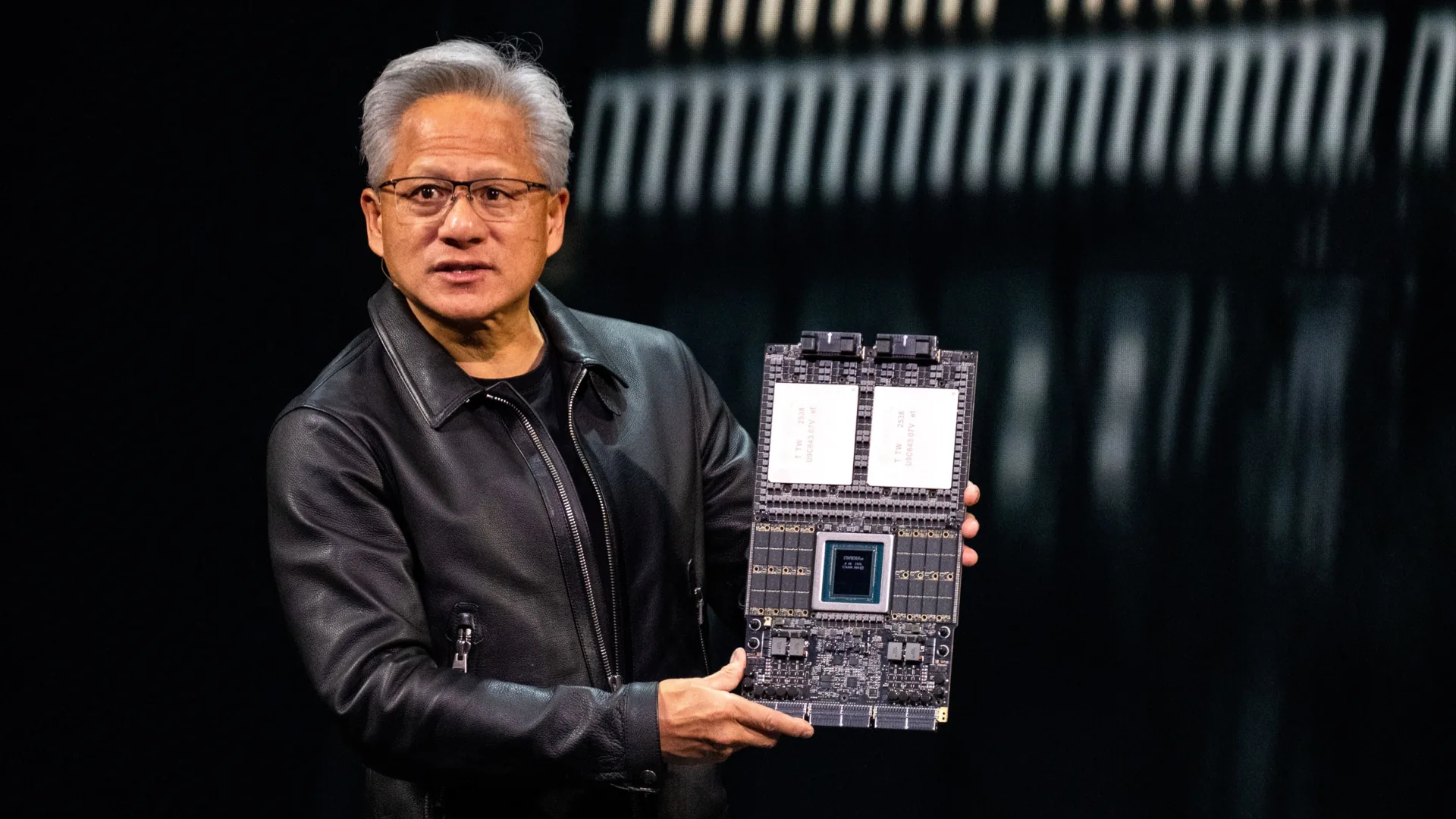 A presenter in a black leather jacket holds a large computing board with visible components and circuitry.