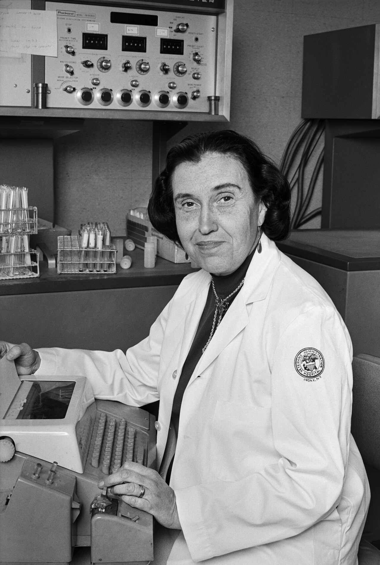 A person in a lab coat is seated at a Packard device with test tubes on the side.