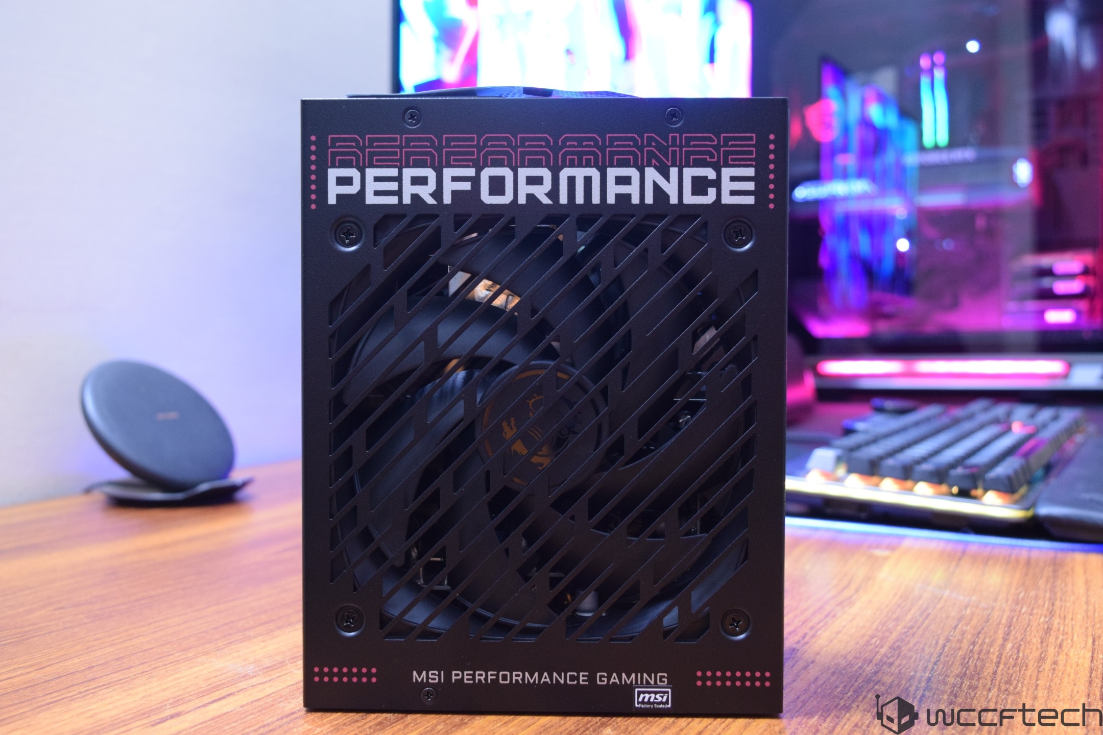 An MSI Performance Gaming power supply unit is displayed on a wooden desk, with the label 'PERFORMANCE' prominently featured on its grille and a colorful gaming setup visible in the background.
